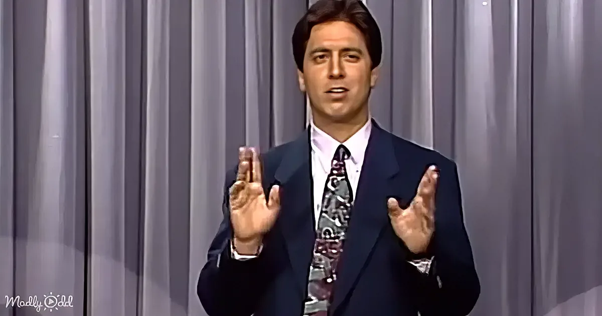 A man in a suit and tie gestures with his hands while speaking, standing against a gray curtain backdrop.