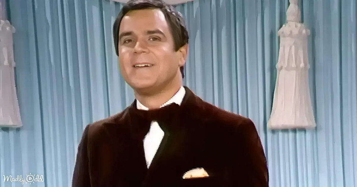 A man in a brown suit and bow tie smiles against a blue curtain backdrop with statues.