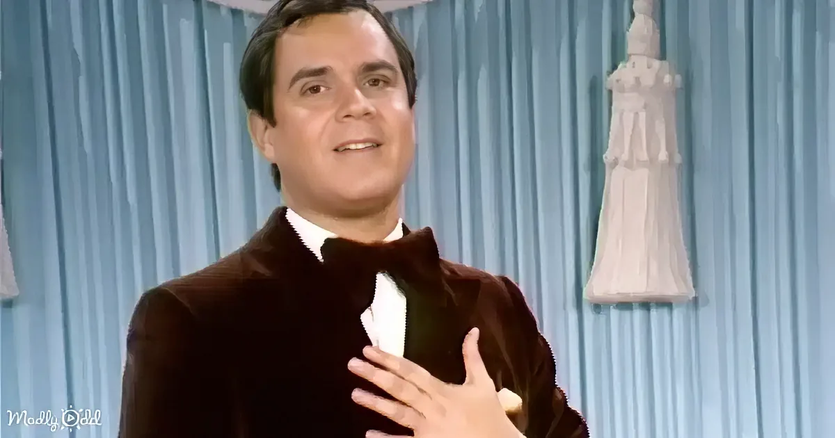 A man in a dark suit gestures with his hand near his chest, standing in front of a blue curtain with a white statue.