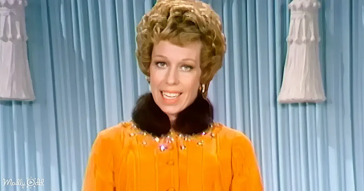 A woman in a bright orange dress with a fur collar smiles against a blue curtain backdrop.
