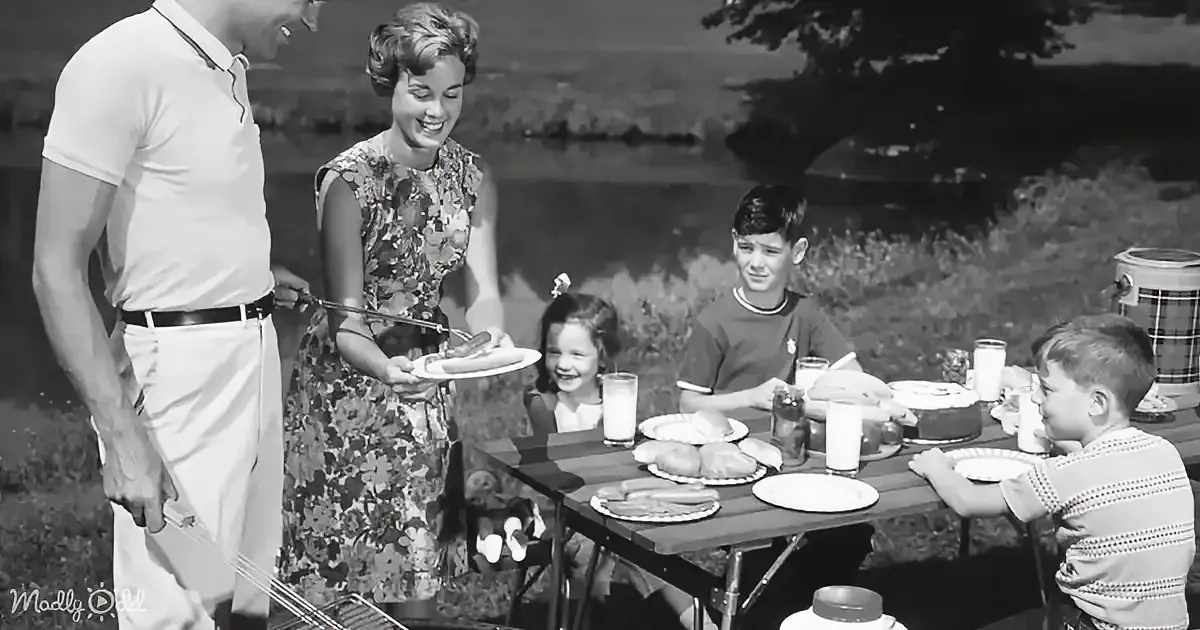 A family enjoys a picnic, with a woman serving food to a man and children seated around a table.