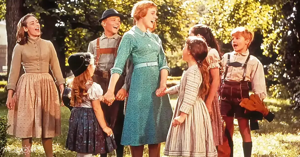 A woman in a blue dress holds hands with a group of people, including children in traditional attire, in a sunny outdoor...