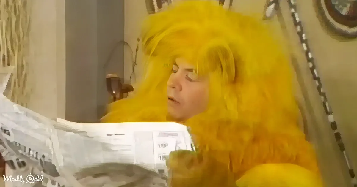 A person with long, bright yellow hair is lying down, holding a glass, and reading a newspaper.