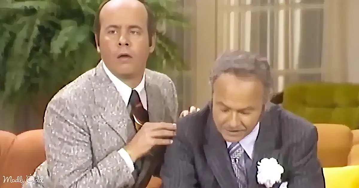 A man in a patterned suit is holding a microphone, while another man in a suit sits beside him, looking down.