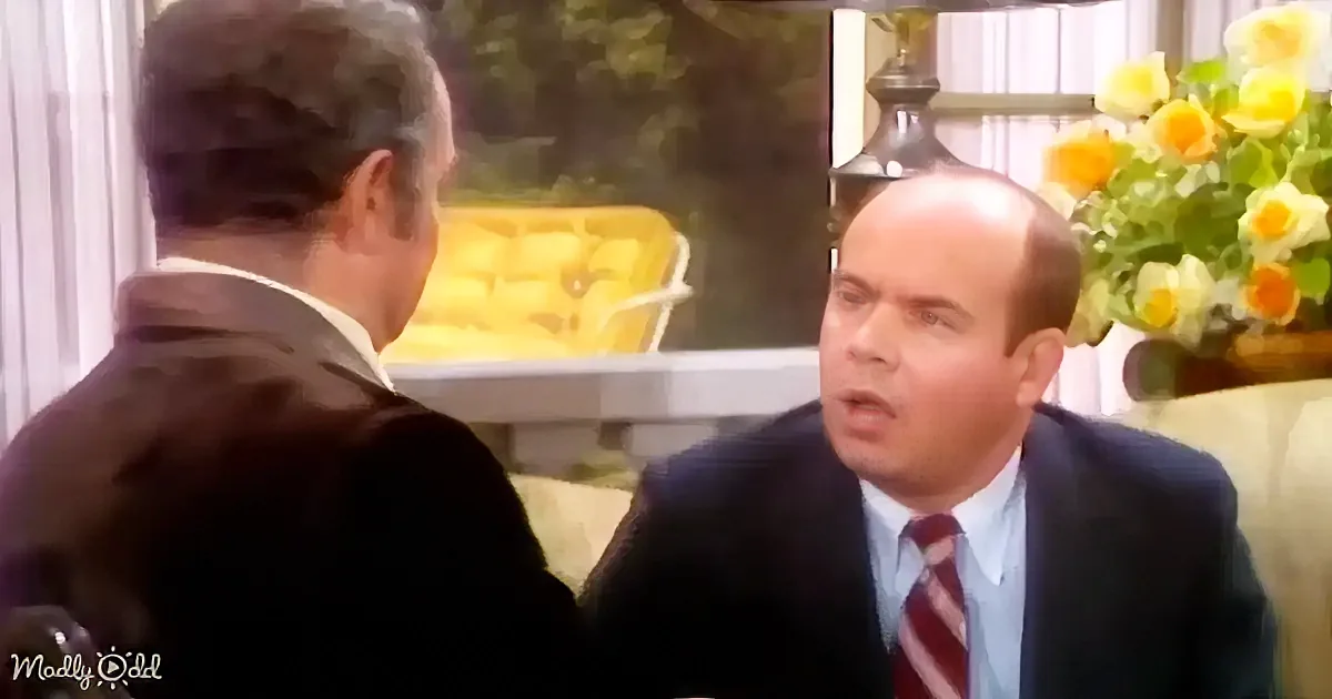 A man in a suit and tie is speaking to another man in a formal setting with flowers and a lamp in the background.