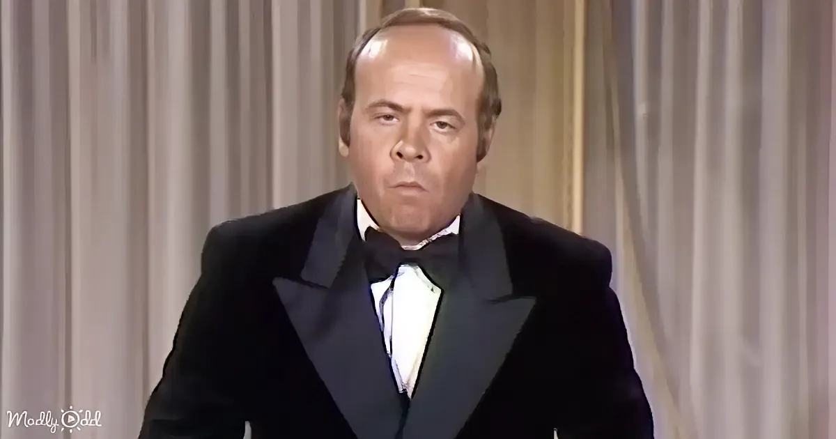 A man in a tuxedo with a bow tie stands against a curtain backdrop.