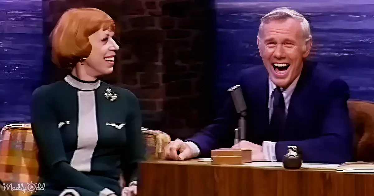 A man and woman are seated at a desk, both smiling and laughing, with a brick wall and microphone in the background.