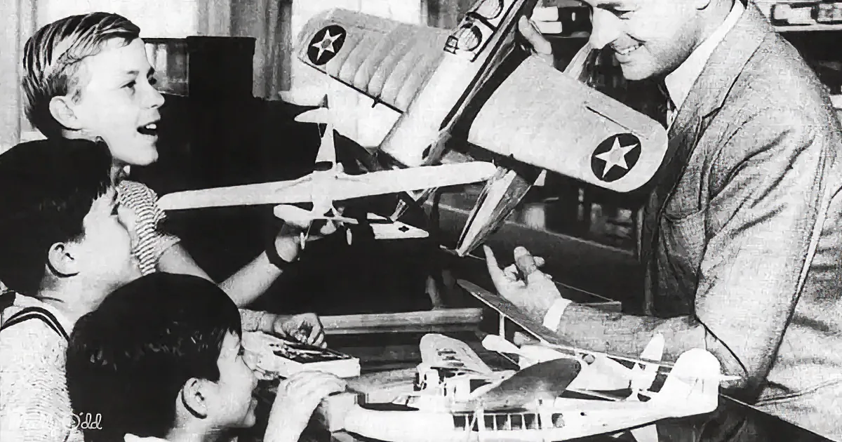 A man and children are assembling model airplanes, with one child holding a plane and another pointing at it.