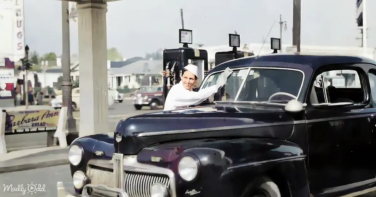 A man in a chef's hat leans out of a vintage car at a gas station, smiling.