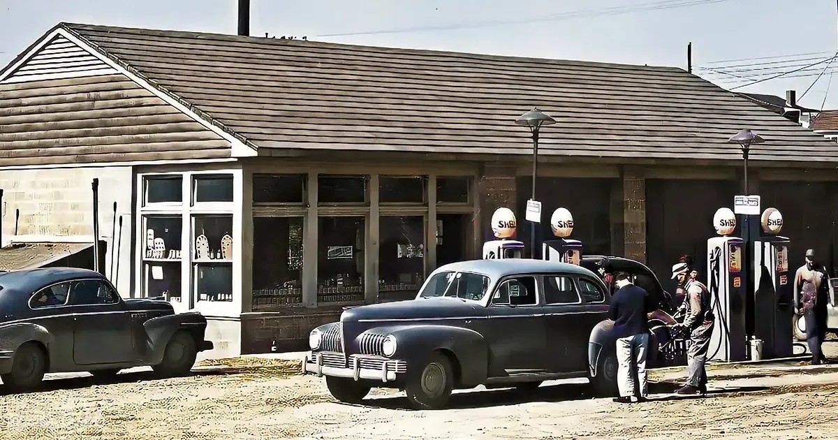 A vintage car is parked at a gas station with people refueling, surrounded by old-style gas pumps and a wooden building.