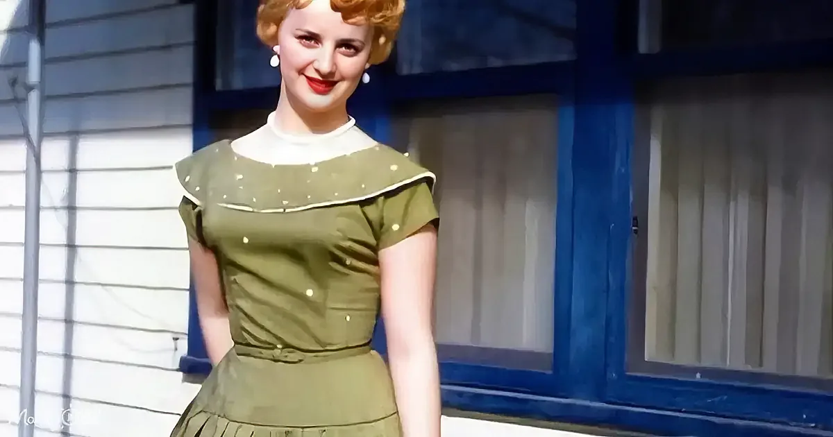 A woman in a green dress with white trim stands in front of a blue-framed window.
