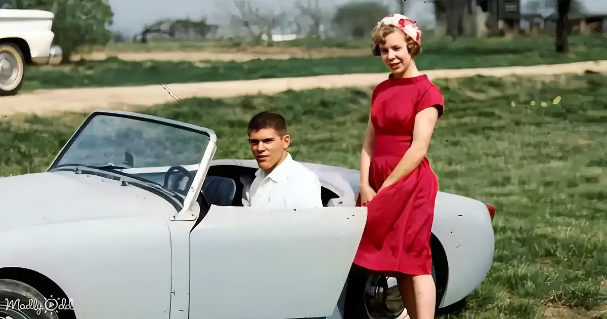 A woman in a red dress stands beside a man in a white car, both smiling in a grassy field.