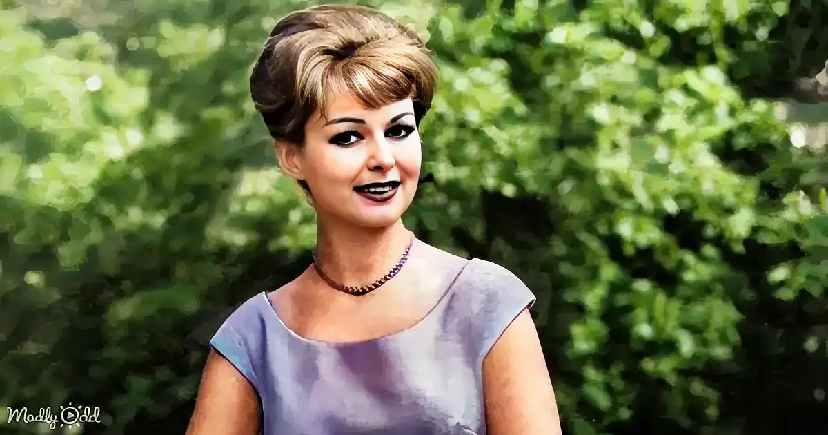 A woman with short, styled hair and makeup, wearing a purple dress and a necklace, stands against a green, leafy background.