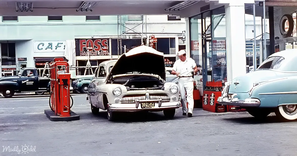 A man in white attire inspects an open hood of a vintage car at a gas station.