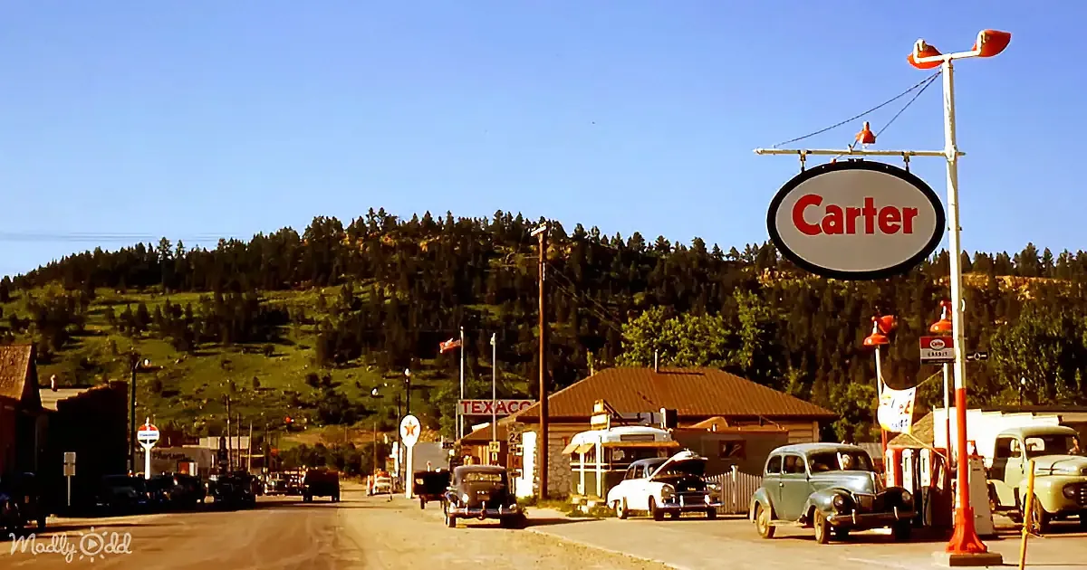 A vintage Carter gas station sign stands tall with classic cars parked nearby, set against a backdrop of green hills and...