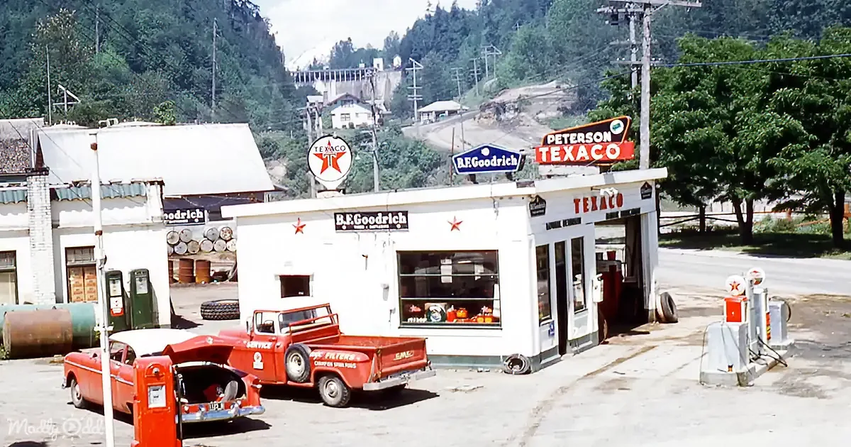 A vintage Texaco gas station with red trucks and classic cars, surrounded by lush greenery and hills.