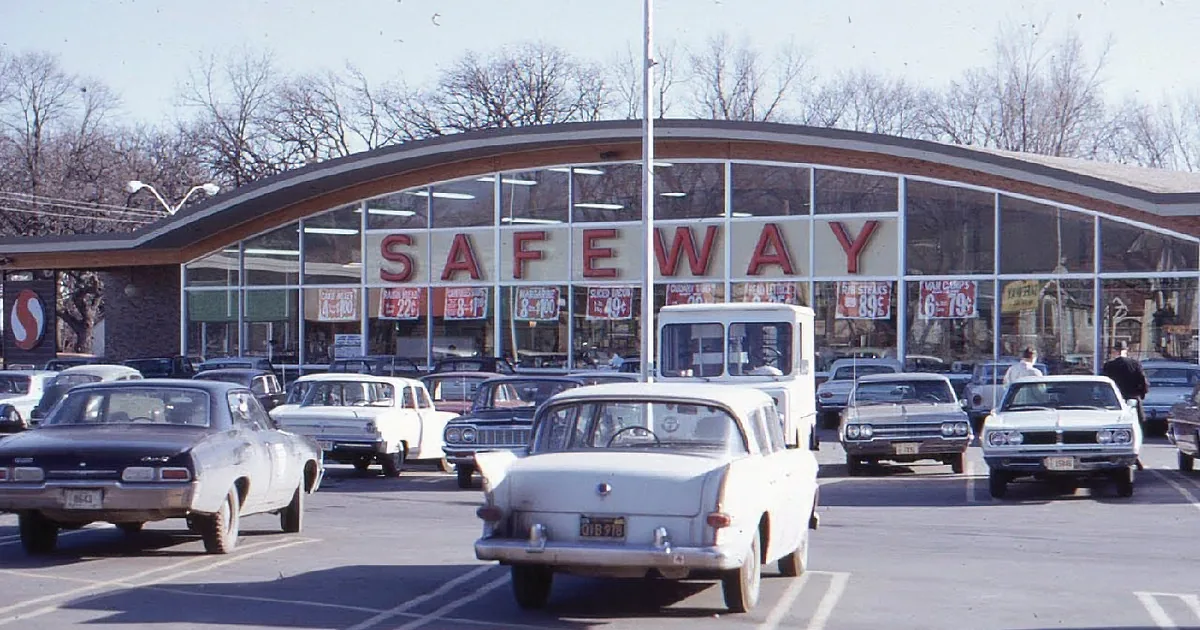 A Safeway store with vintage cars parked in front, featuring classic 1960s vehicles.