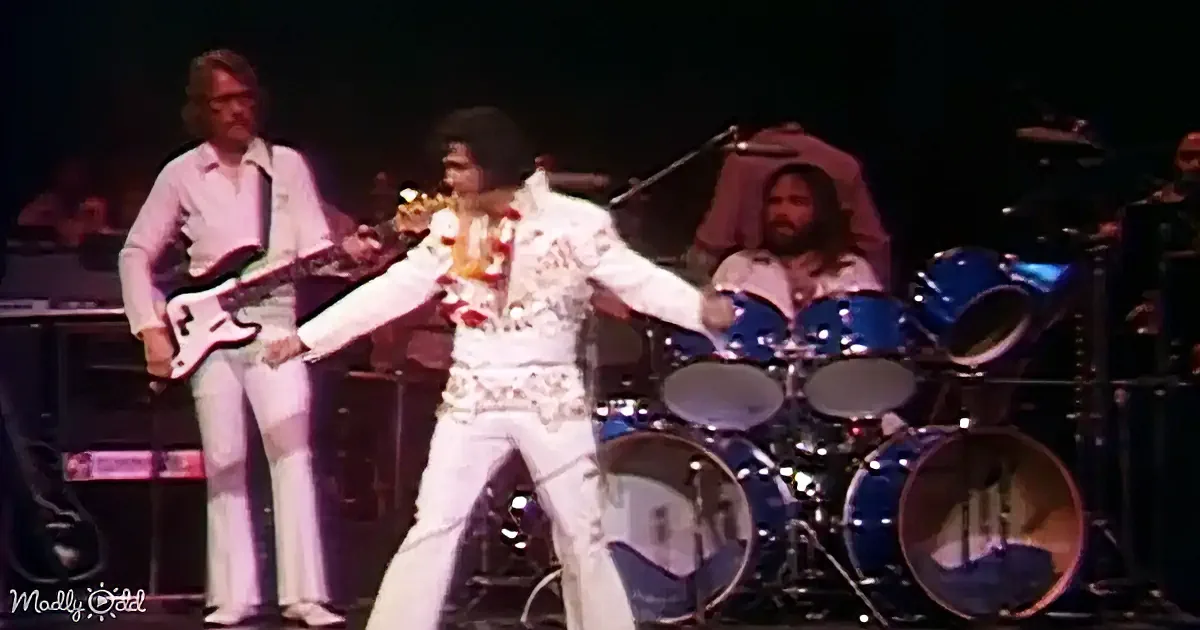 Elvis Presley in white jumpsuit dances on stage with band, including guitarist and drummer.