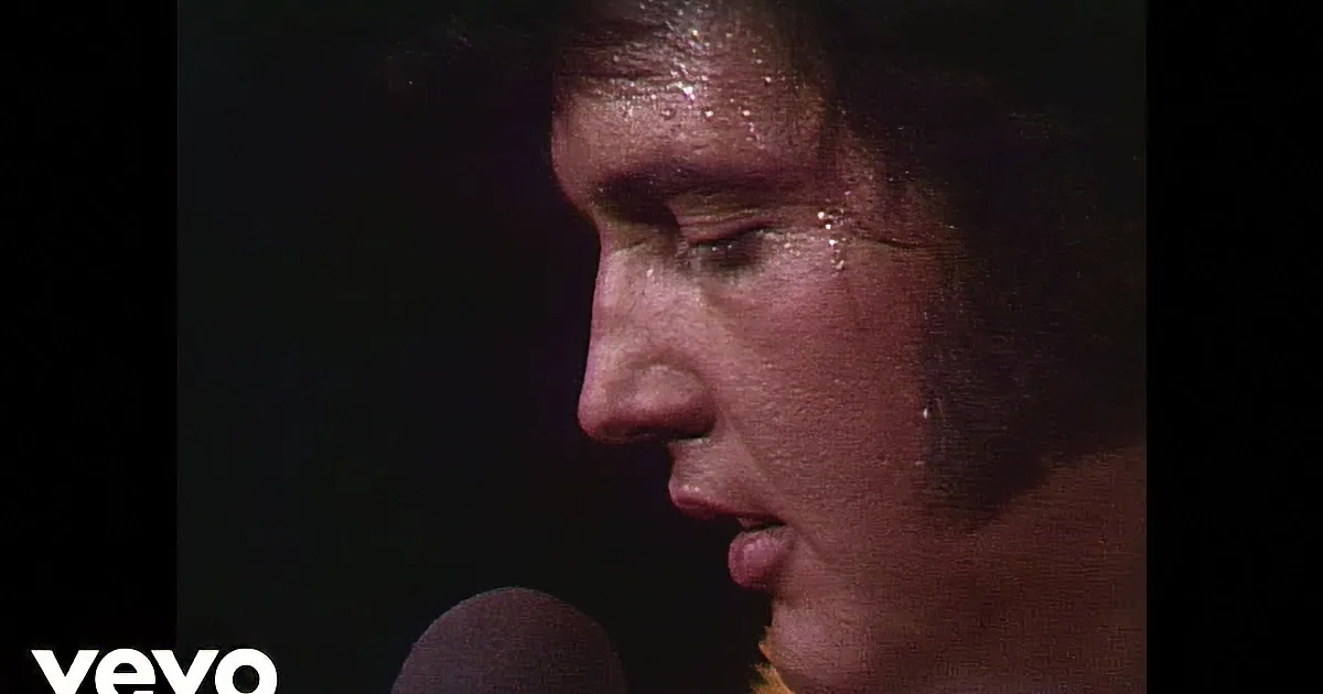 Elvis Presley in a white sequined jumpsuit with a microphone, surrounded by a yellow lei.