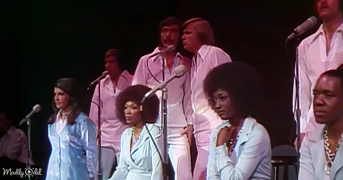 A group of performers in white and blue outfits sing on stage, with microphones and a dark background.
