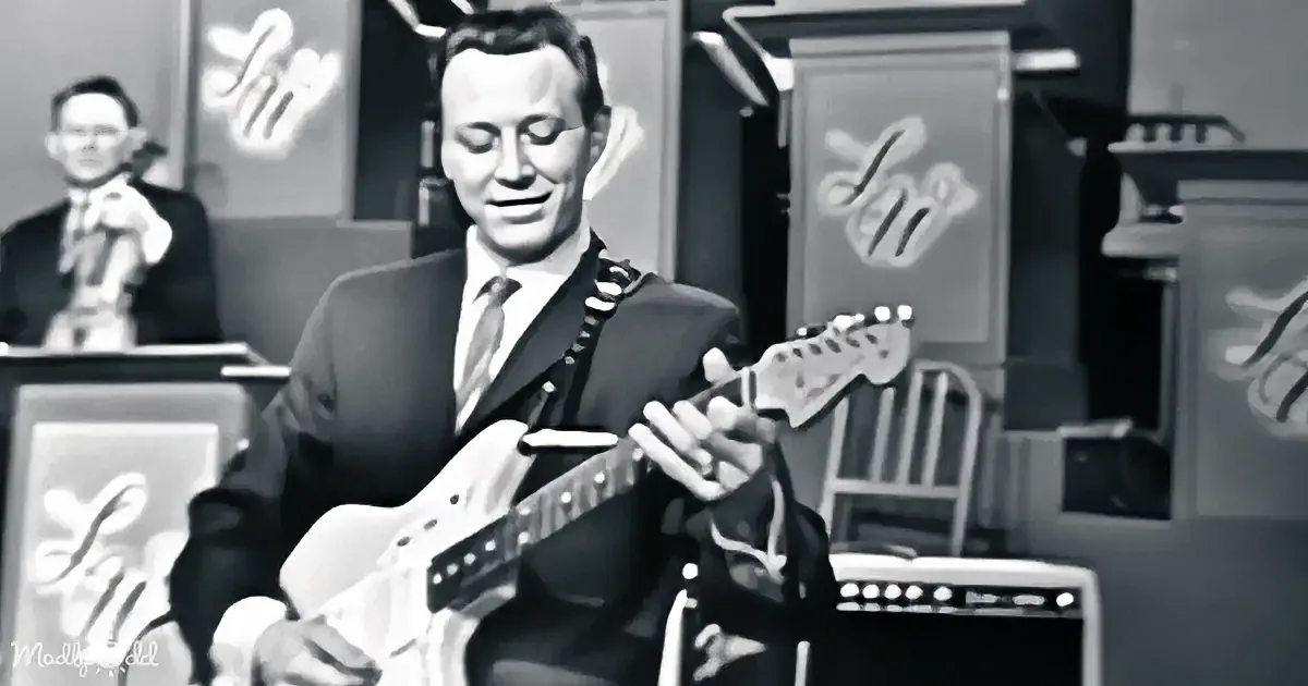 A man in a suit plays an electric guitar on stage, with a violinist in the background.