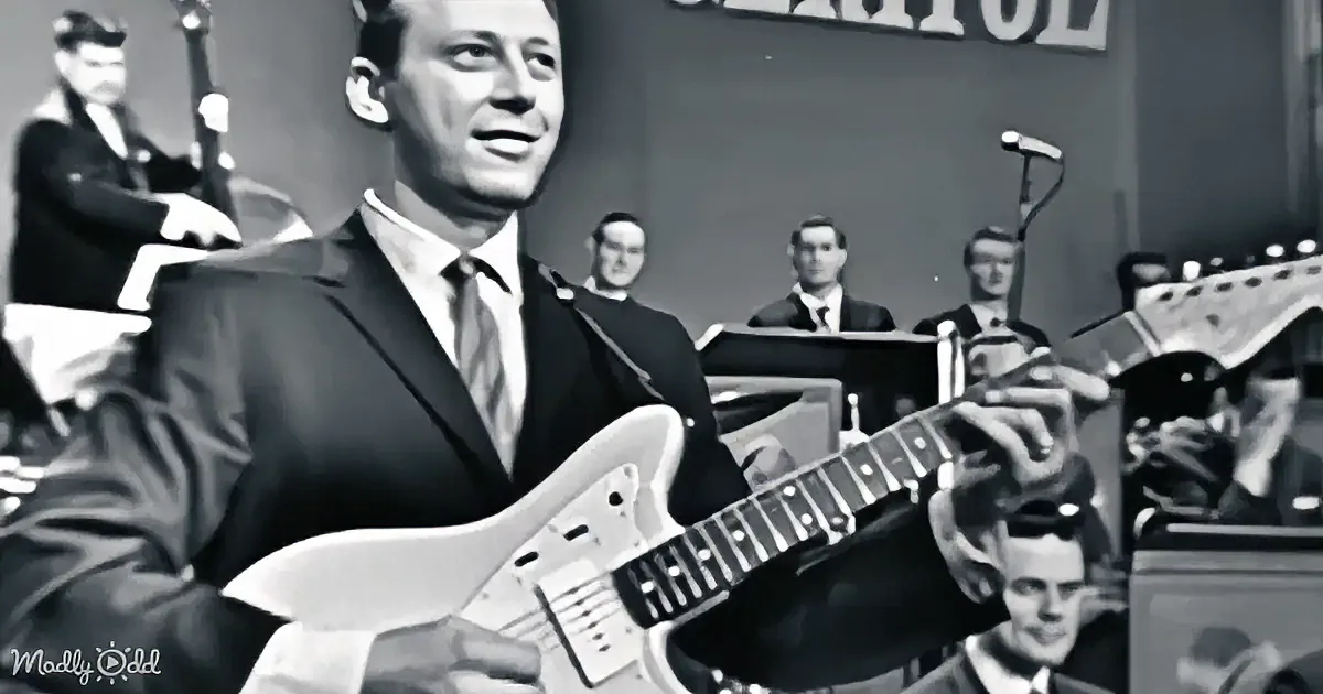 A man in a suit plays a guitar on stage with a band behind him.