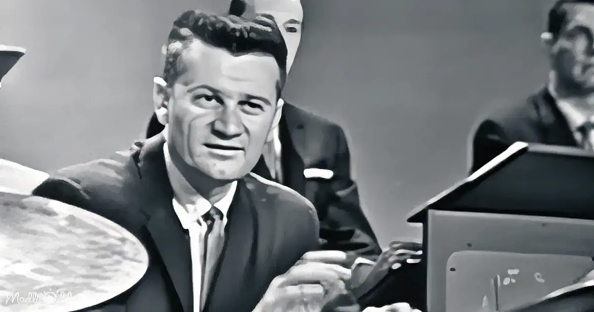 A man in a suit and tie sits at a desk, smiling, with a drum set and other musicians in the background.