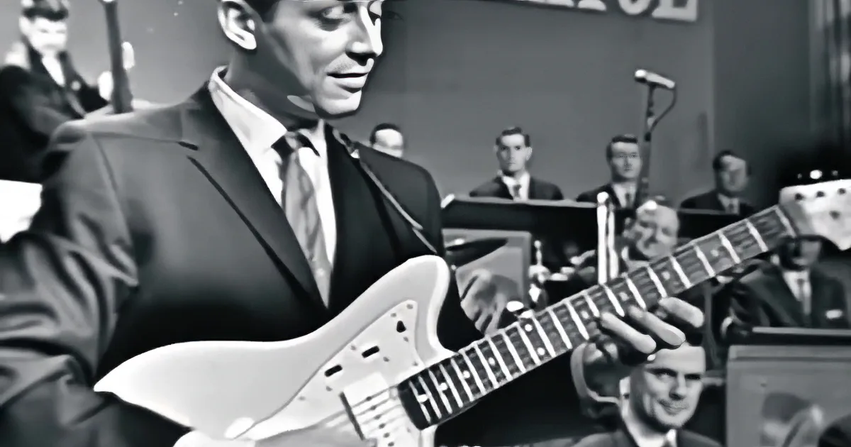 A man in a suit and tie smiles while playing a white electric guitar.