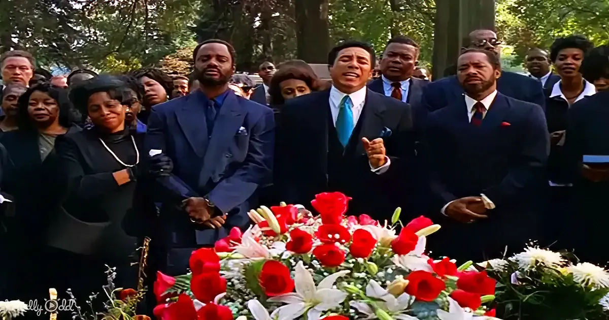A man in a suit stands at a floral arrangement, surrounded by a group of people in formal attire.