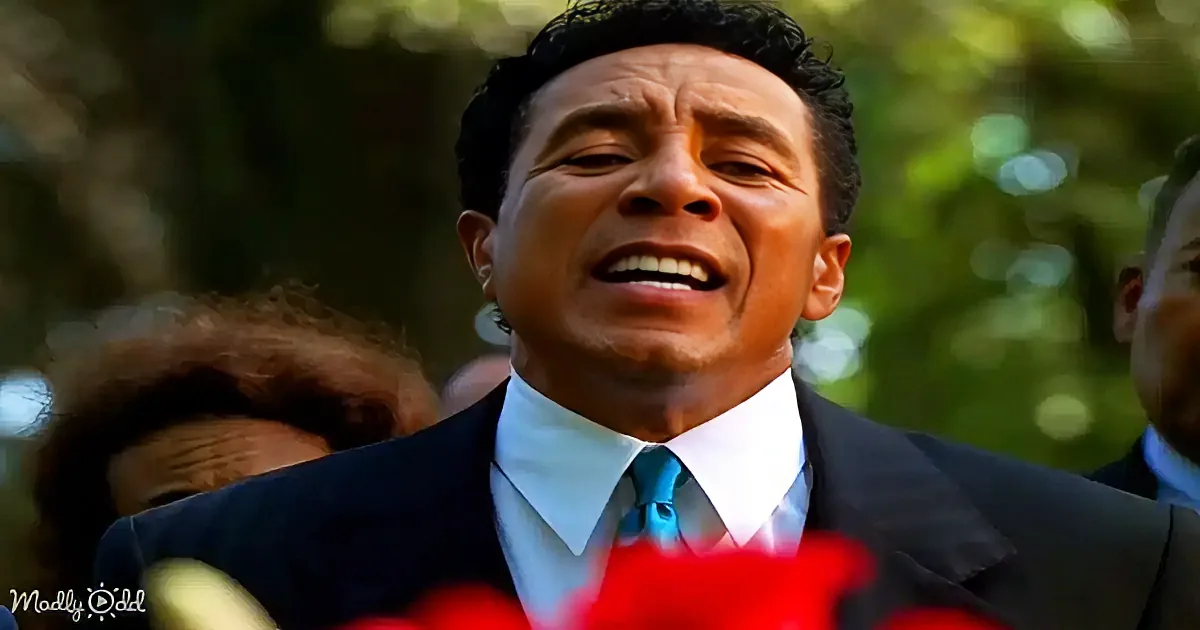 A man in a suit and tie is singing passionately, surrounded by blurred figures and greenery.