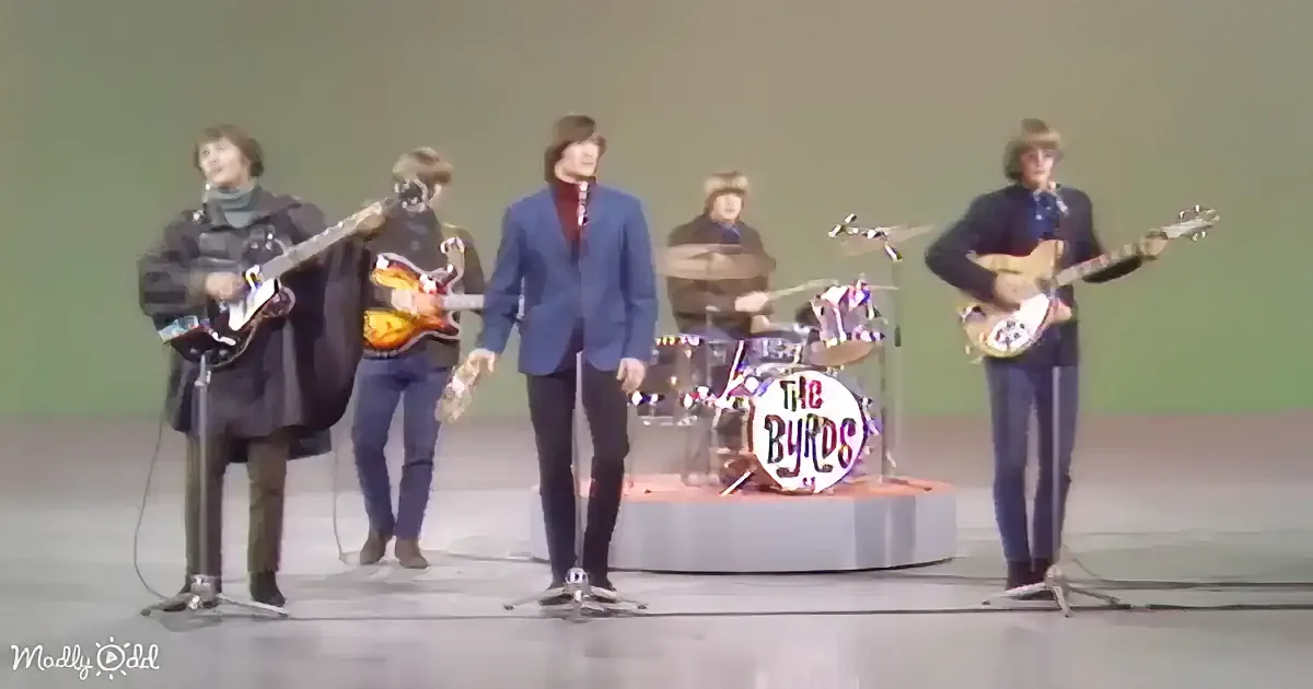 The Byrds perform on stage with guitars and drums, dressed in 1960s attire.