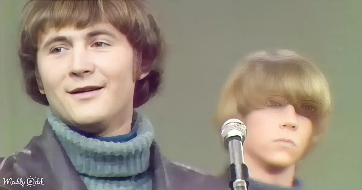 A smiling person with shoulder-length hair, wearing a sweater, stands in front of a microphone.