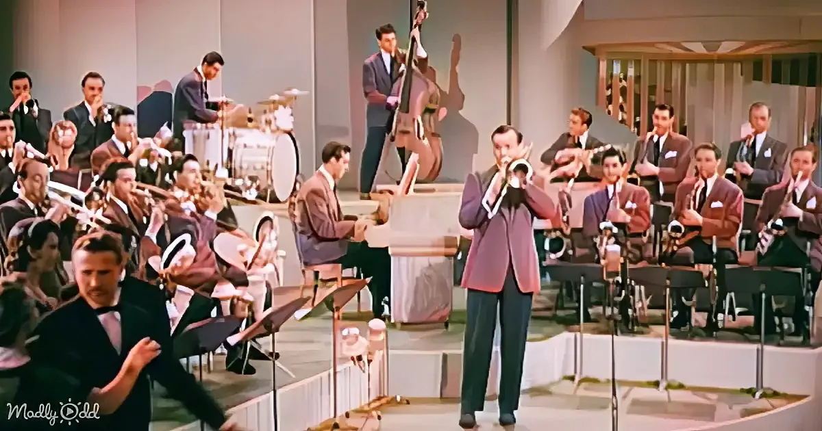 A man in a suit plays a trumpet in front of a large orchestra on stage.