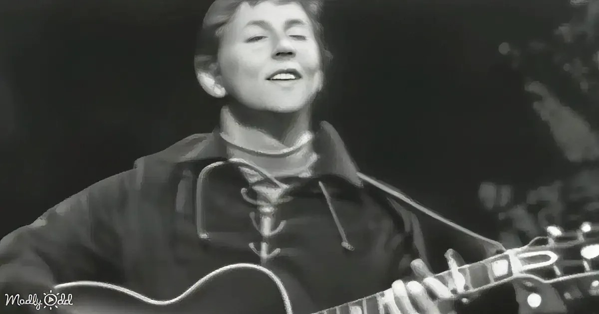 A person in a dark jacket and turtleneck plays an acoustic guitar, smiling with eyes closed.