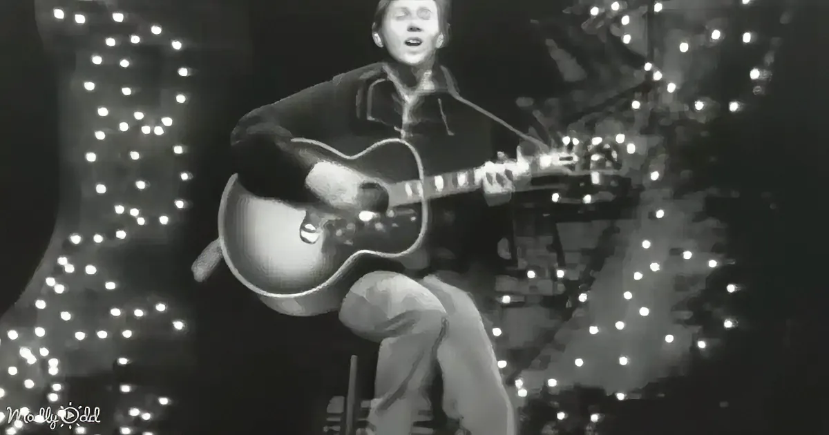 A person in a dark outfit plays a guitar on stage with bright lights in the background.