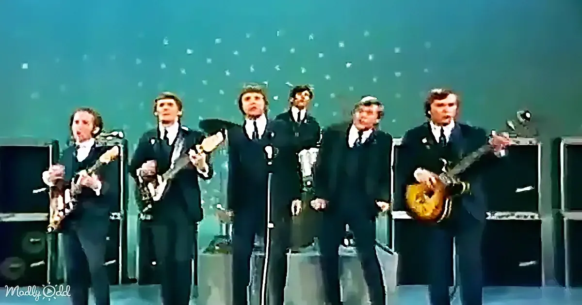 A group of six men in suits perform on stage, holding guitars and singing into microphones.