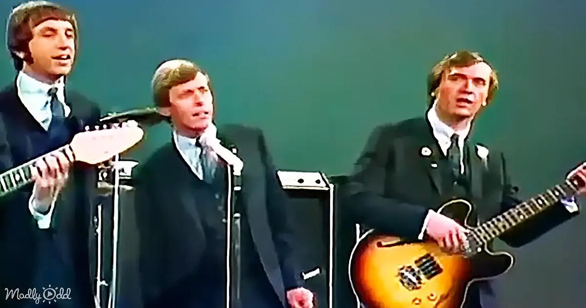 Three men in suits sing and play guitars on stage, one holding a white guitar and another a yellow one.