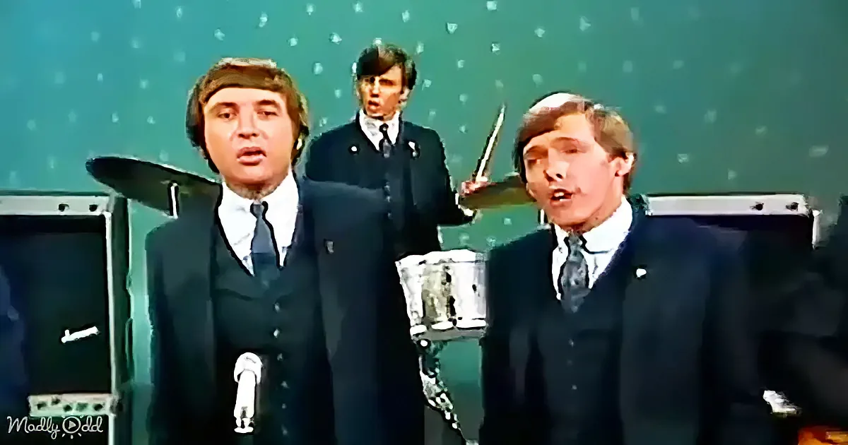 Three men in suits sing into microphones, one plays drums behind them.