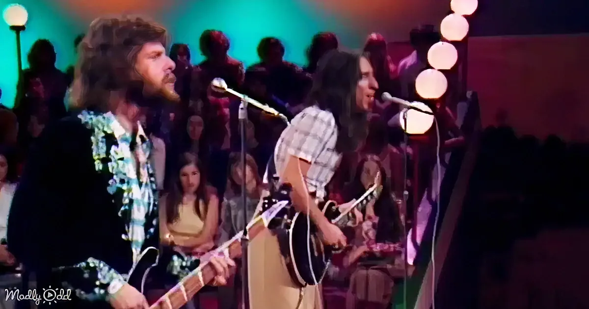 Two musicians with long hair and guitars perform on stage, surrounded by a crowd and colorful lights.