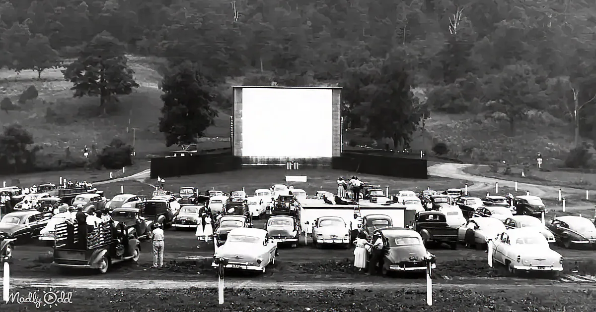 A vintage drive-in movie theater with cars parked in rows, people mingling, and a large screen in the background.