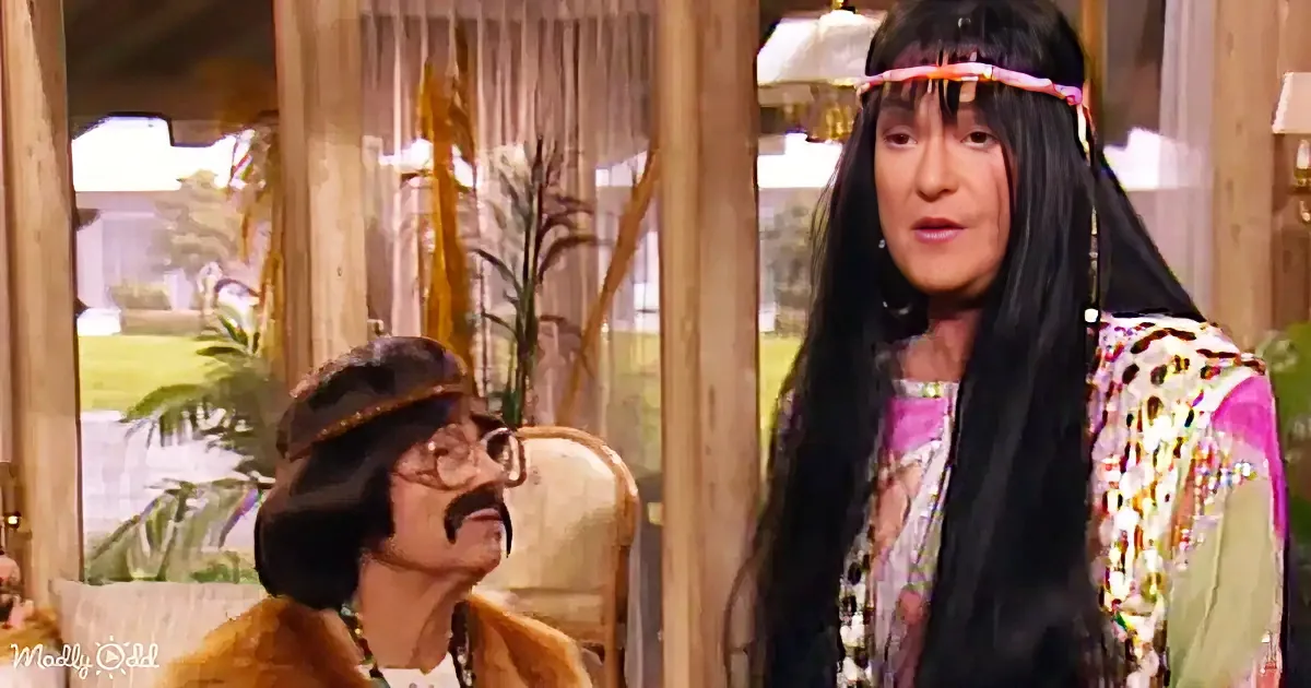 A woman with long black hair and a colorful outfit stands next to a man in a brown jacket and glasses, both in a room...