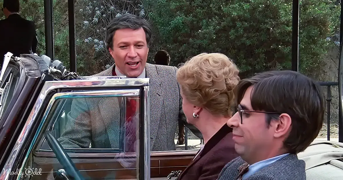 A man in a suit leans into a car, speaking to two passengers inside, with trees and a person in the background.