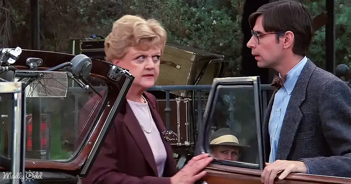 A woman in a maroon suit and pearl necklace stands beside a man in a suit, both near an old car with a child inside.