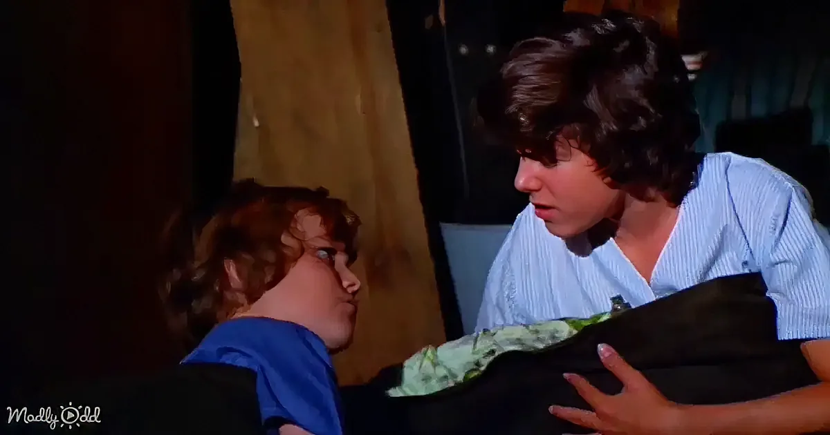 A woman in a striped shirt leans over a bed, engaging in a conversation with a child in a blue shirt.