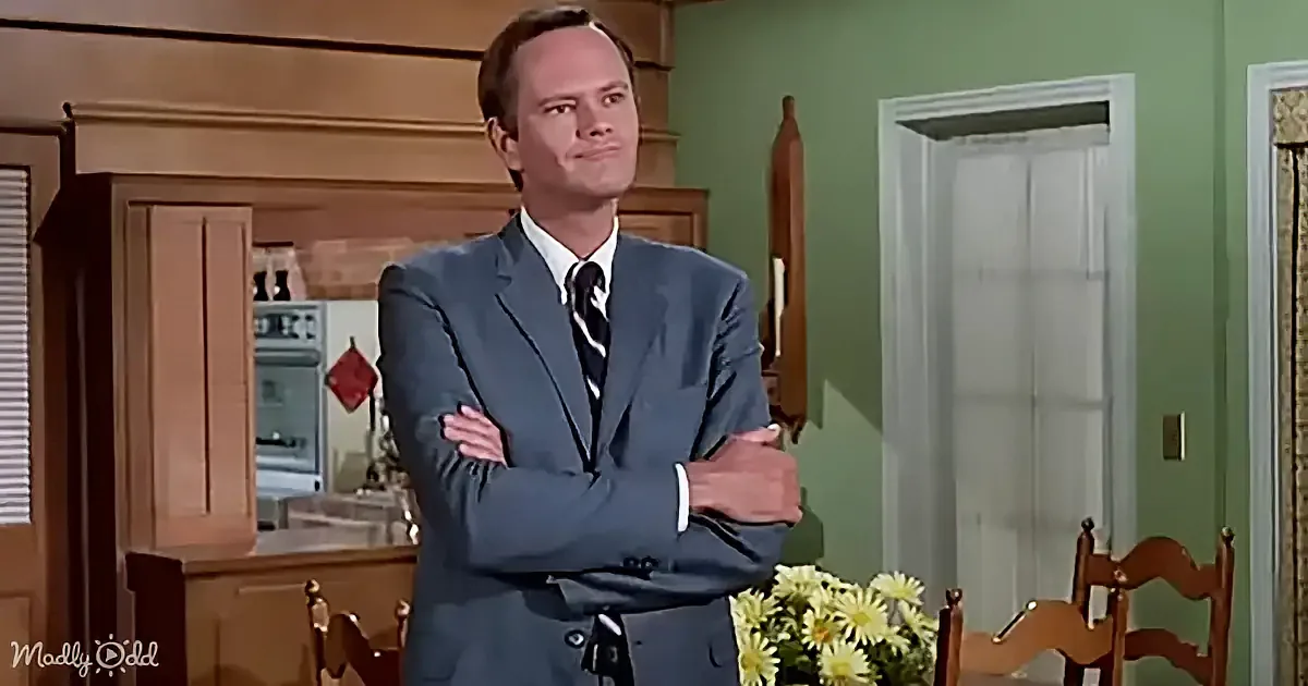 A man in a gray suit stands with arms crossed in a kitchen, looking to the side.