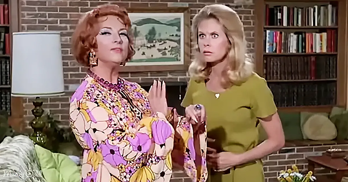 A woman in a floral dress gestures while another in a green dress looks surprised in a living room setting.
