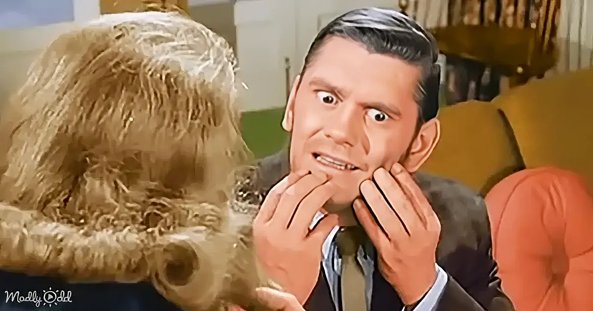 A man in a suit holds his face in a surprised or shocked expression, looking at a woman with long blonde hair.