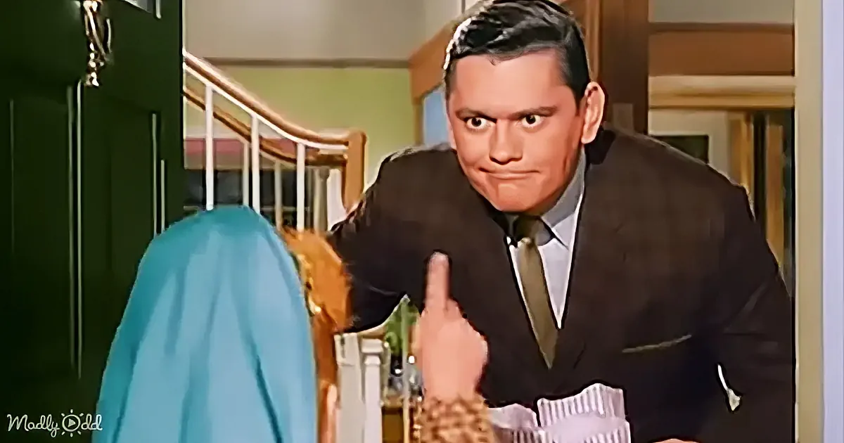 A man in a suit points upwards while holding a newspaper, with a staircase and green door in the background.