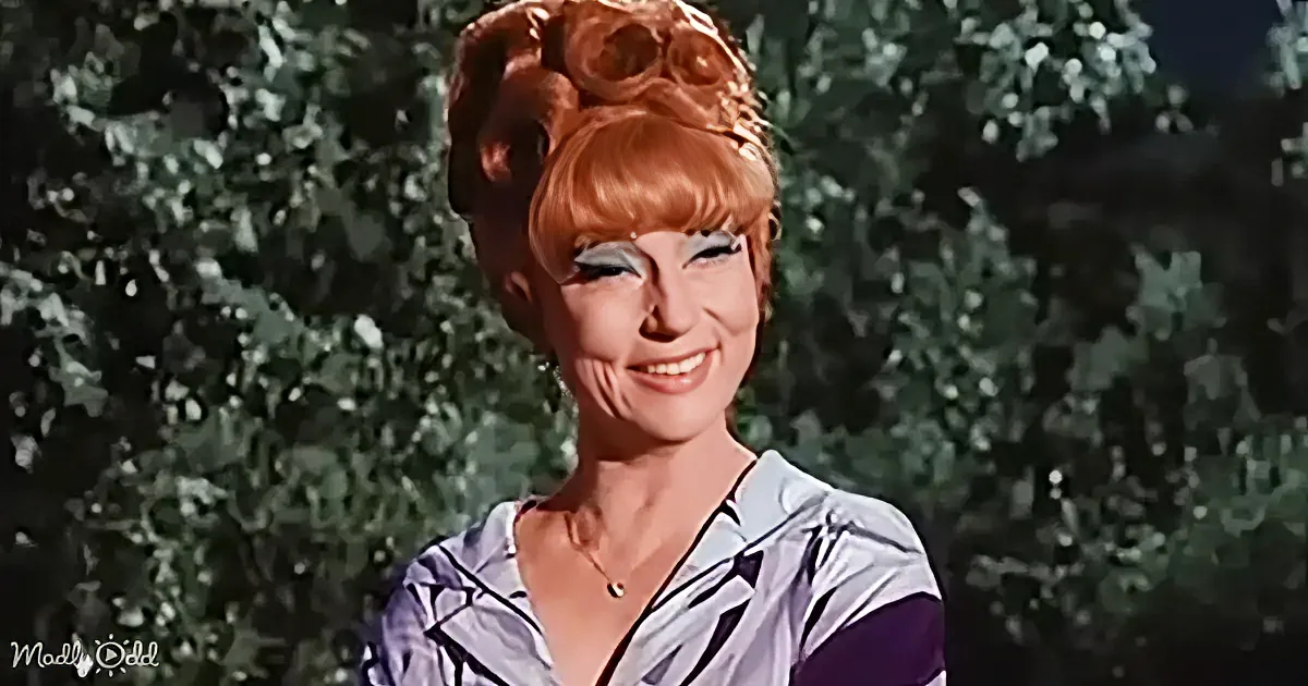 A smiling woman with red hair styled in a bun, wearing a patterned top and necklace, stands outdoors with trees in the...