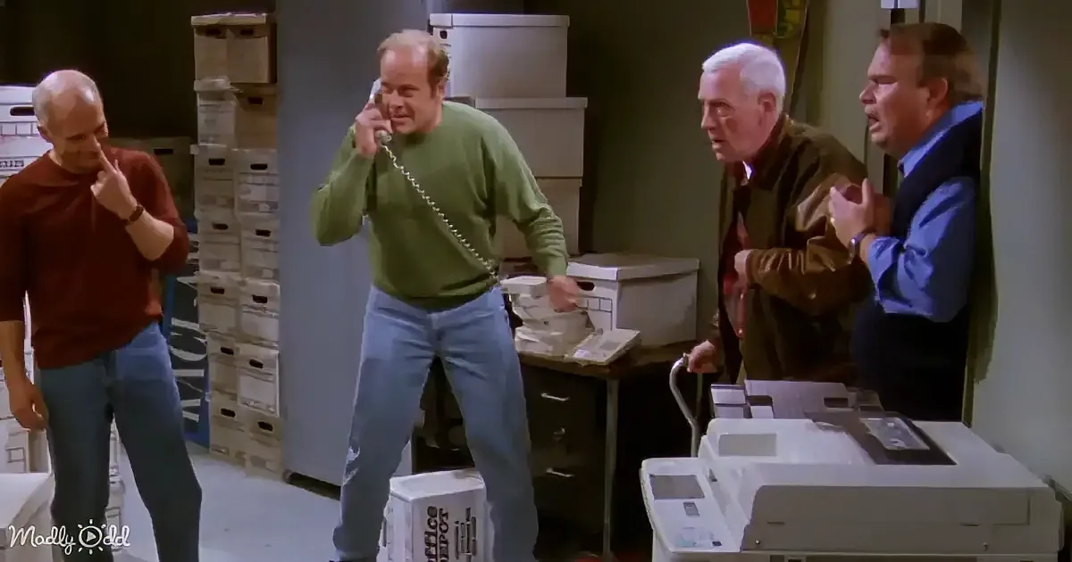 A man in a green sweater is on the phone, surrounded by others in a cluttered office space.